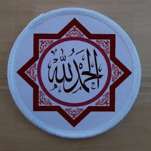 Puede incluir: Parche redondo blanco con un diseño de estrella roja de ocho puntas. La estrella tiene un círculo rosa en el centro con la palabra árabe "Allah" escrita en caligrafía negra.