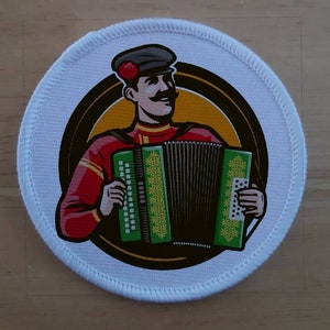 May include: A white round patch with a colorful illustration of a person playing an accordion. The person is wearing a red and black outfit with a black hat and a red flower. The accordion is green, black, and yellow with floral designs.