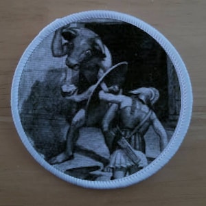 May include: A black and white embroidered patch depicting a scene of a warrior fighting a mythical creature. The warrior is holding a shield and a sword, while the creature has large horns and a muscular body.