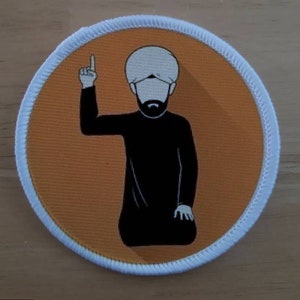 Puede incluir: Un parche ilustrado en blanco y negro de una persona que lleva un turbante blanco y ropa negra con el brazo levantado, apuntando hacia arriba. El parche está sobre un fondo marrón con un borde blanco.