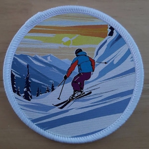 Könnte beinhalten: Runder Aufnäher mit einem Skifahrer in einer blauen Jacke und lila Hose, der einen schneebedeckten Berg hinunterfährt. Der Hintergrund zeigt einen Sonnenuntergang über schneebedeckten Bergen und immergrünen Bäumen. Der Aufnäher hat einen weißen Rand.