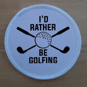 Puede incluir: Parche redondo blanco con texto negro y un diseño de bola de golf y palos. El texto dice "I'd Rather Be Golfing".