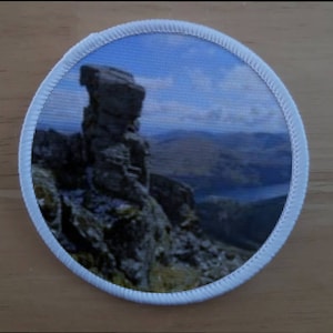 May include: A round white patch with a printed image of a rocky mountain peak against a blue sky with white clouds. The mountain peak is a jagged rock formation.