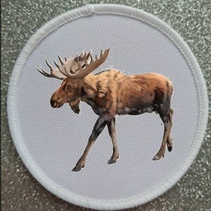 May include: A white round patch with a brown and white moose illustration. The moose is walking to the right with its head turned to the left.