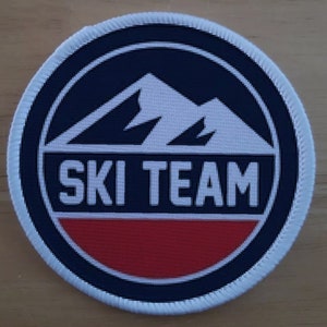 May include: White and red embroidered patch with a black circle and white text that reads "SKI TEAM". Inside the circle is a white mountain range on a dark blue background.
