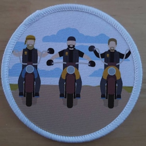 Può includere: Tre personaggi dei cartoni animati che guidano motociclette su una strada marrone chiaro con un cielo blu e nuvole bianche sullo sfondo. I personaggi indossano giacche e caschi gialli e marroni. Le motociclette sono marroni e nere.