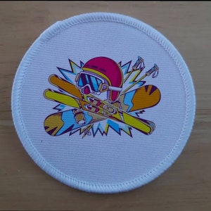 May include: A circular white patch with a skiing-themed design. The illustration shows a skier's helmet, goggles, skis, and poles in vibrant colors, set against a backdrop of yellow and blue lightning. The patch has a white, stitched edge.
