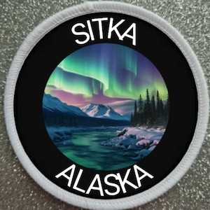 May include: A round white patch with a black circle in the center. The black circle has a white border and the text "SITKA ALASKA" in white. Inside the black circle is a photo of a river flowing through a snowy landscape with a green and purple aurora borealis in the sky.