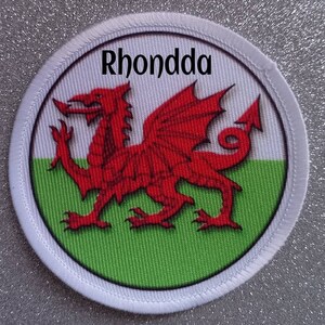 Può includere: Una toppa circolare bianca con un drago rosso su uno sfondo verde e bianco. Il testo "Rhondda" è stampato in nero nella parte superiore della toppa.