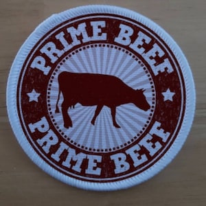 Op de afbeelding: Een ronde witte patch met een rode rand en een rood silhouet van een koe in het midden. De tekst "PRIME BEEF" staat in een rode cirkelvormige rand rond de koe.