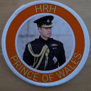 Könnte beinhalten: Ein runder weißer und orangefarbener Aufnäher mit dem Text "HRH PRINCE OF WALES" in goldenen Buchstaben. Die Mitte des Aufnähers zeigt ein Foto eines Mannes in Militäruniform.