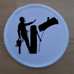 May include: White round patch featuring a black silhouette of a person climbing a tree with a chainsaw. The person is wearing a helmet and harness. The tree has a branch that has been cut off.