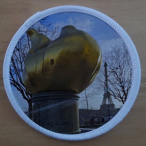 May include: A gold-colored sculpture of a flame, with the Eiffel Tower visible in the background. The sculpture is on a pedestal and is surrounded by trees.