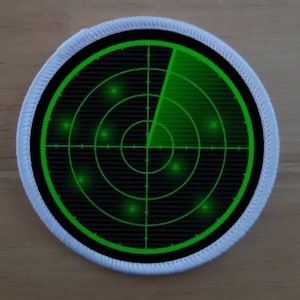 May include: A white circular patch with a black border and a green radar screen inside. The radar screen has a green line extending from the center to the edge of the screen. There are several green dots on the screen.
