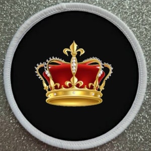 Puede incluir: Una corona dorada con joyas rojas y perlas blancas sobre un fondo negro. La corona está rodeada por un borde blanco.