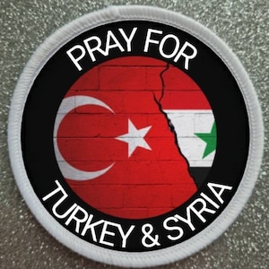 May include: A white circular patch with black text that reads "Pray for Turkey & Syria". The center of the patch features a brick wall with the Turkish and Syrian flags painted on it, separated by a crack.