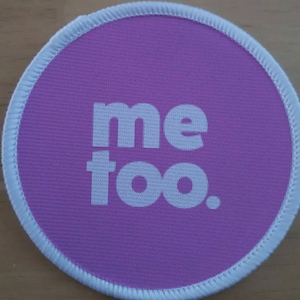 Op de afbeelding: Een ronde, paarse stoffen badge met witte stiksels rond de rand. De tekst "me too." is in wit op de badge gedrukt.