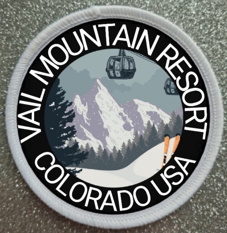 May include: A white round patch with black text and a graphic of a snowy mountain scene with ski lifts and skiers. The text reads "Vail Mountain Resort Colorado USA".