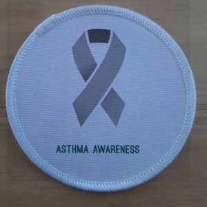 Op de afbeelding: Een ronde witte stoffen patch met een grijs lint en de tekst "ASTHMA AWARENESS" in groen.