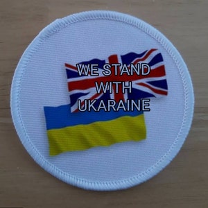 May include: White round patch with the text "WE STAND WITH UKRAINE" and the flags of the United Kingdom and Ukraine.