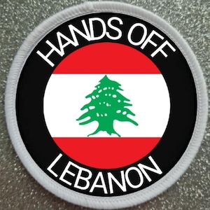 Może przedstawiać: Biała okrągła łatka z czarnym tekstem, który brzmi "Hands Off Lebanon" wokół czarnego koła z libańską flagą w środku. Flaga jest czerwona, biała i zielona z zielonym cedrem w środku.