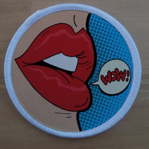 Peut inclure: Un patch de style bande dessinée rouge et blanc avec les lèvres d'une femme disant "WOW!" dans une bulle de dialogue. L'arrière-plan est bleu avec des pois blancs.