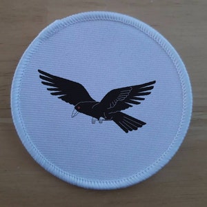 May include: A white round patch with a black crow flying in the centre. The crow has a red eye.