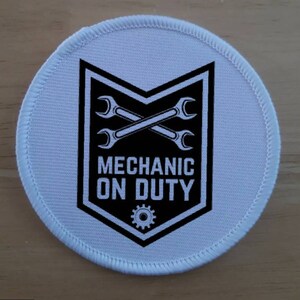May include: A round, white patch with a black and white graphic. The graphic features crossed wrenches inside a black shield, with the words "MECHANIC ON DUTY" below. A gear is at the bottom of the shield. The patch has a white stitched border.