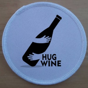 Puede incluir: Parche redondo de tela blanca con una silueta negra de una botella de vino que está siendo abrazada por dos manos. El texto "HUG WINE" está impreso debajo de la botella.