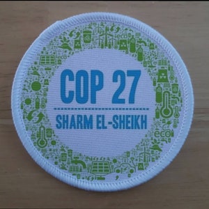 May include: A white circular patch with a green border. The border is filled with green icons related to climate change. The centre of the patch has the text "COP 27" in blue and "SHARM EL-SHEIKH" in blue below.