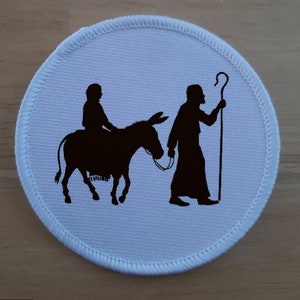 May include: A white round patch with a black silhouette of a person riding a donkey and another person walking behind them holding a staff.