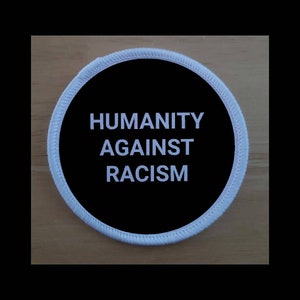 Può includere: Toppa rotonda con la scritta bianca "HUMANITY AGAINST RACISM" su sfondo nero. La toppa ha un bordo bianco cucito ed è posta su una superficie di legno marrone chiaro.