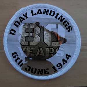 Peut inclure: Un écusson rond blanc avec du texte noir qui dit "D-Day Landings 30 Years 6th June 1944". L'écusson présente une photo d'un casque militaire avec un motif de camouflage.