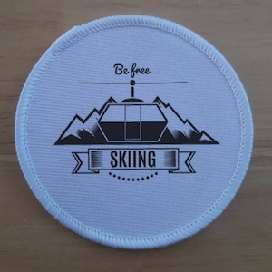 Puede incluir: Parche redondo blanco con un gráfico en blanco y negro de un telesilla en las montañas. El texto "Be free" está por encima del telesilla y el texto "SKIING" está por debajo del telesilla.