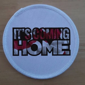 May include: White round fabric patch with the text "It's Coming Home" printed in black with a red, white, and blue design behind the text.