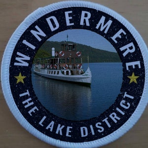 May include: A white and blue round patch with the text "Windermere The Lake District" and a picture of a boat on a lake.
