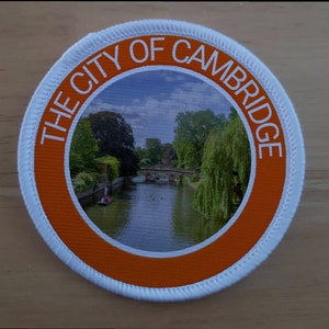 Op de afbeelding: Een witte ronde patch met een oranje rand. De rand heeft de tekst "THE CITY OF CAMBRIDGE" in wit. Het midden van de patch toont een foto van een rivier met een brug en bomen.