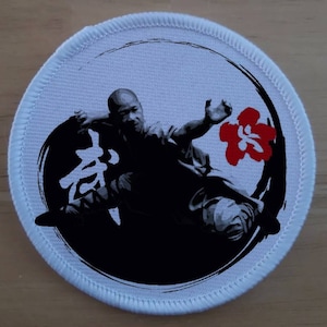 May include: A white round patch with a black and white image of a person in a martial arts pose. The person is surrounded by a black circle with a red Chinese character in the center. The character is surrounded by a red flower.