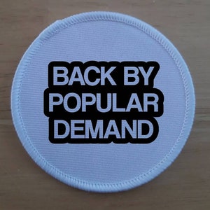 Puede incluir: Parche redondo de tela blanca con texto negro que dice "Back by Popular Demand".