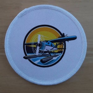 May include: A white round patch featuring a colourful image of a seaplane flying over a sunset with palm trees. The seaplane is blue and white with a yellow propeller. The sunset is orange and yellow with a blue sky. The palm trees are green with brown trunks.