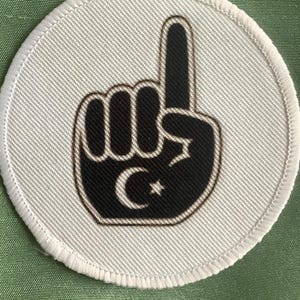 Puede incluir: Parche circular blanco con una imagen en blanco y negro de una mano con el dedo índice apuntando hacia arriba. La mano sostiene una luna creciente negra y una estrella.