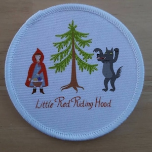 Peut inclure: Un écusson rond en tissu blanc avec une fille en capuchon rouge portant un panier, un pin vert et un loup gris aux yeux jaunes. Le texte "Little Red Riding Hood" est imprimé en rouge sous l'image.