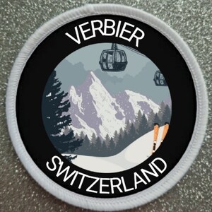 May include: A white and black circular patch with the text "VERBIER SWITZERLAND" in white lettering. The patch features a black and white illustration of a mountain range with a ski lift and skiers.