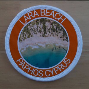 Puede incluir: Un parche redondo blanco y naranja con el texto "Lara Beach Paphos Cyprus" y una foto de una playa con agua turquesa.