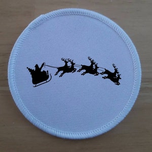 Può includere: Toppa rotonda in tessuto bianco con una silhouette nera di Babbo Natale su una slitta trainata da renne.
