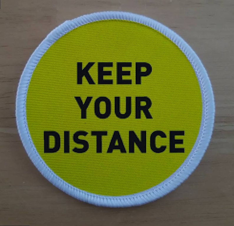 Puede incluir: Un parche redondo de tela amarillo con un borde blanco. El texto "KEEP YOUR DISTANCE" est&aacute; impreso en negro.