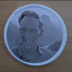 May include: A black and white photograph of a man smiling, printed on a white circular patch with a white stitched border.