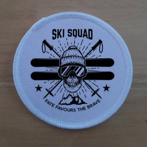 May include: White round patch with black and white graphic design. The graphic features a skull wearing a ski mask and goggles, two crossed ski poles, and the text "SKI SQUAD" and "FATE FAVOURS THE BRAVE".
