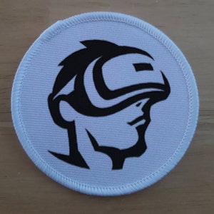 Può includere: Una toppa rotonda bianca con una grafica nera di un profilo umano stilizzato che indossa una visiera o un casco futuristico. La toppa ha un bordo bianco cucito, probabilmente un emblema o un logo.