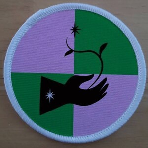 May include: A round patch with a white border and a green and pink checkered background. A black silhouette of a hand holding a small plant with a star on the stem is in the centre of the patch.